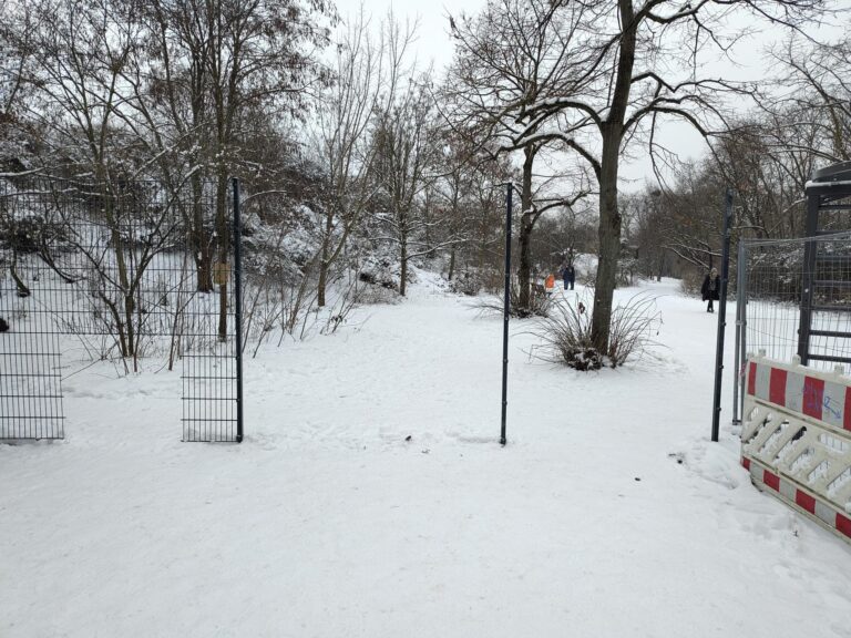 Schneebedeckter Weg im Görlitzer Park mit offenstehendem Zauntor und Absperrgittern, im Hintergrund spazierende Menschen.