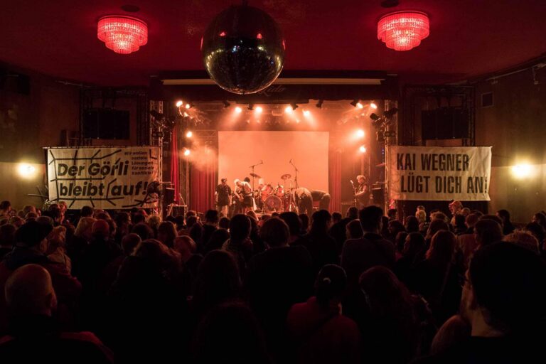 Konzertsaal mit Publikum vor einer Bühne, daneben Banner „Der Görli bleibt auf!“ und „Kai Wegner lügt dich an!“