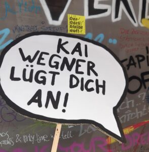 Sprechblasen-Schild 'Kai Wegner lügt dich an!' vor einer beschmierten Wand mit Aufkleber 'Der Görli bleibt auf'