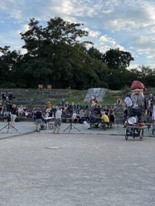 Menschen sitzen bei einer Kiezversammlung auf den Treppenstufen im Görlitzer Park, daneben eine große Zwergenfigur mit Schild „Görli bleibt auf!“.