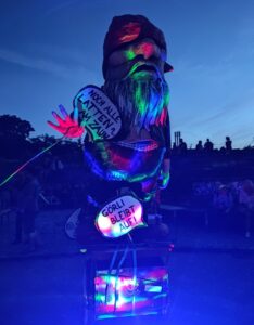 Bunt beleuchtete Gartenzwerg-Figur bei Nacht mit Schildern „Noch alle Latten am Zaun?“ und „Görli bleibt auf!“