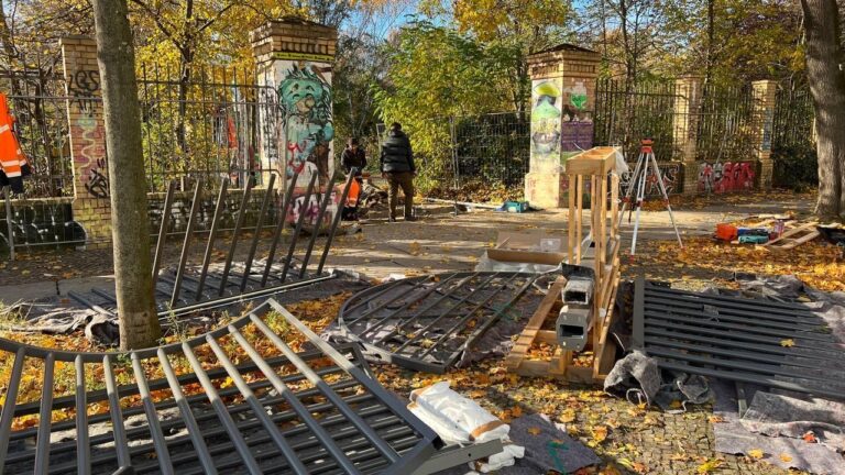 Baustelle im Görlitzer Park: Zerlegte Zaunelemente liegen auf dem Boden, im Hintergrund arbeiten zwei Personen an einem mit Graffiti besprühten Torpfeiler.