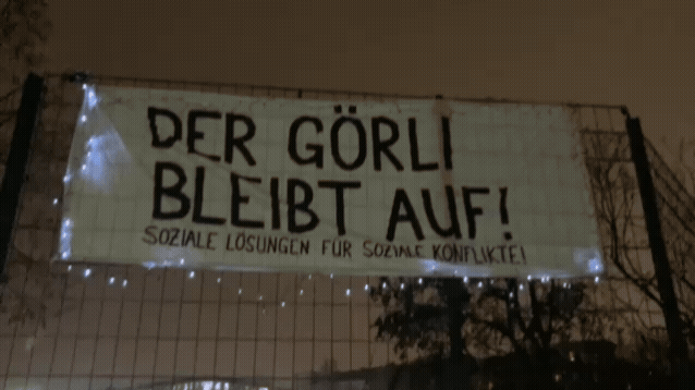 Banner „Der Görli bleibt auf! Soziale Lösungen für soziale Konflikte!“ nachts an einem Zaun, umrahmt von einer Lichterkette.