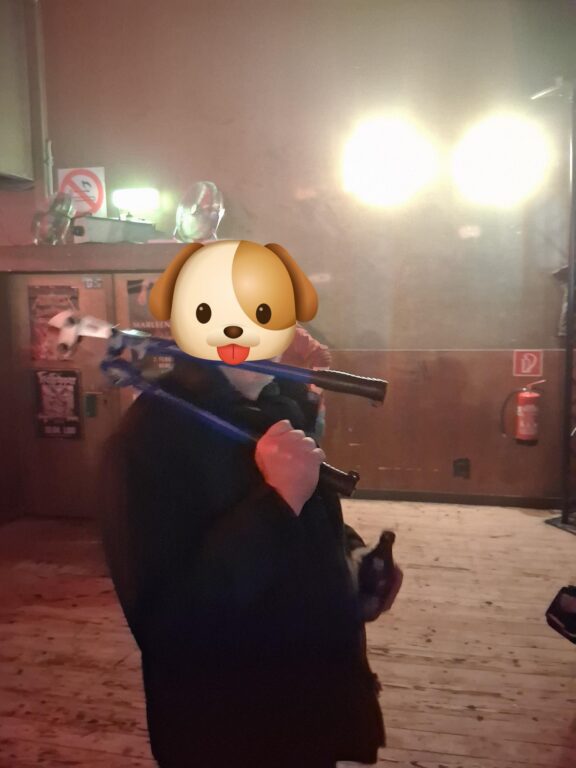 Person mit Trommelstöcken in dunkler Kleidung in einem Veranstaltungsraum, Gesicht durch ein Hunde-Emoji verdeckt.