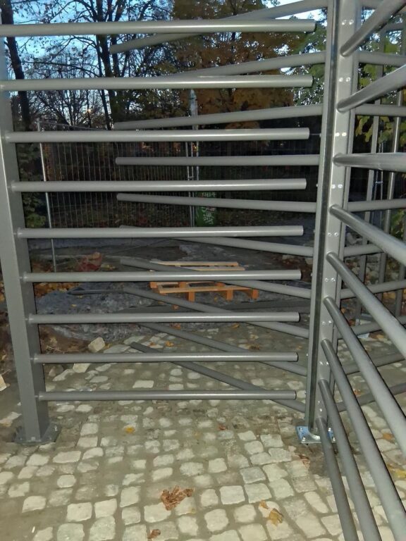 Neu installiertes Drehkreuz-Tor aus grauen Metallstreben an einem Parkeingang, im Hintergrund Bäume.