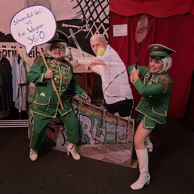 Zwei Personen in grünen Uniformen posieren mit Tennisschlägern neben einer Pappfigur von Kai Wegner, Schild 'Schwarz-Rot Görli vs. Kai Wegner 36:0'