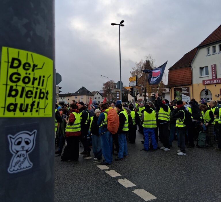 Menschenmenge in gelben Warnwesten bei einer Kundgebung; im Vordergrund ein Sticker „Der Görli bleibt auf!“ an einem Mast.