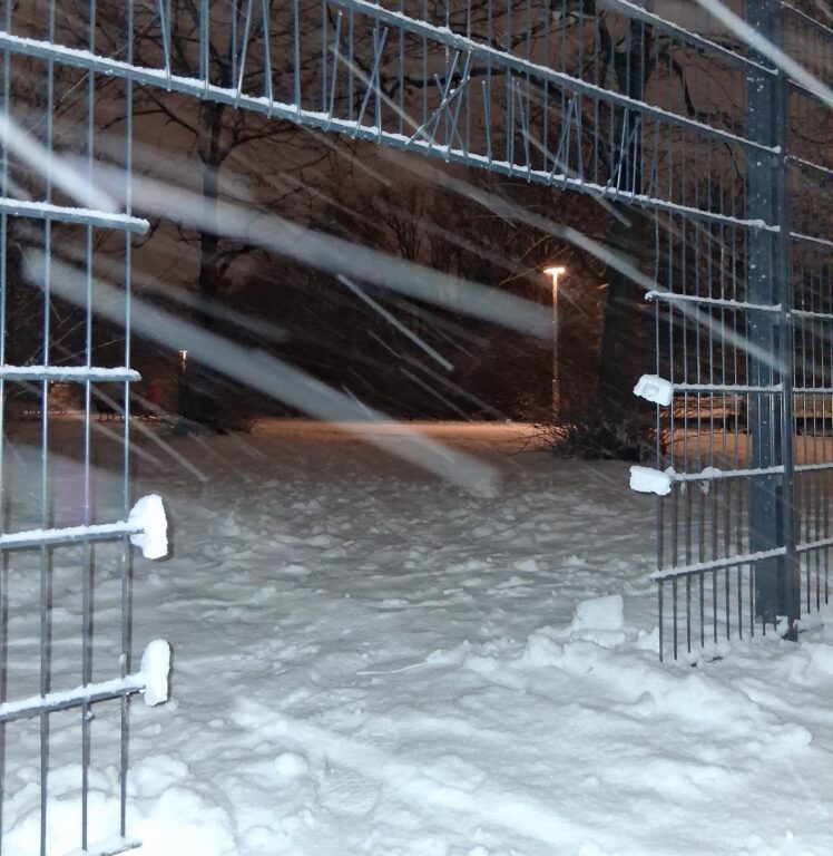 Nächtliche Schneeszene: Lücke zwischen zwei Zaunelementen am Görlitzer Park, dahinter ein verschneiter, beleuchteter Weg.
