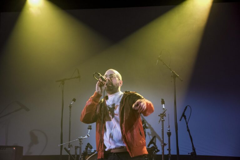 Sänger mit Brille und oranger Jacke performt auf einer Bühne im Scheinwerferlicht