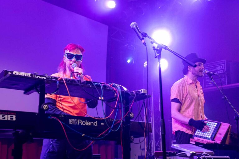 Zwei Musiker auf einer Bühne mit violettem Bühnenlicht; einer singt an Keyboards mit Aufschrift „Quentz“, der andere bedient einen Sampler.