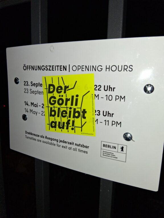 Nächtliches Foto eines Öffnungszeiten-Schilds am Görlitzer Park, überklebt mit gelbem Aufkleber „Der Görli bleibt auf!“.