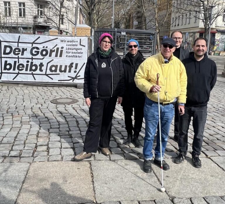 Fünf Personen stehen vor einem Banner 'Der Görli bleibt auf!' auf dem Kopfsteinpflaster, einer nutzt einen Blindenstock