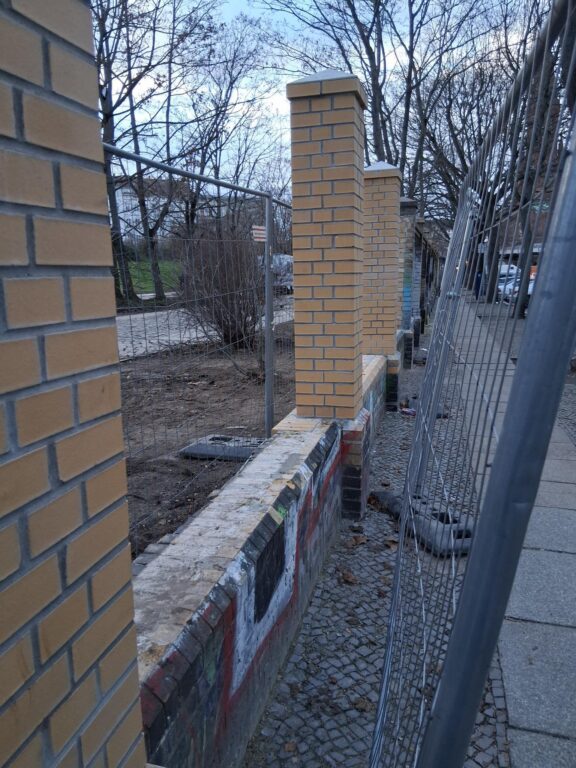 Backsteinmauer mit neuen Mauerpfeilern und Metallzaun entlang eines Gehwegs, im Hintergrund kahle Bäume.