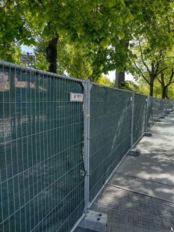 Grüner Bauzaun mit Sichtschutz entlang eines Gehwegs unter Bäumen, mit Firmenschild „tetris“.