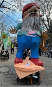 Überlebensgroße Zwergenfigur mit rotem Hut und blauer Latzhose bei einer Demonstration, im Hintergrund weitere Teilnehmende.